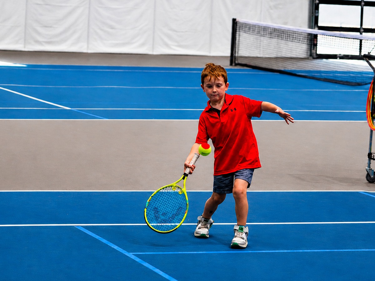 Young boy hitting a forehand with a red ball during a junior tennis lesson on the indoor courts at RacFit in Buda, Texas