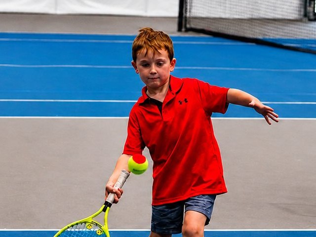 Young boy hitting a forehand with a red training ball beside a ball cart during a Red Ball junior tennis class at RacFit in Buda, Texas